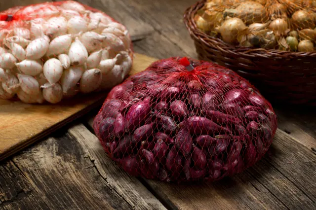 Červená cibule v síťovém pytlíku vedle česneku na dřevěném povrchu.